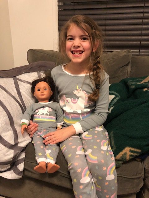  Young girl in unicorn PJs smiling at camera while holding a doll in matching PJs